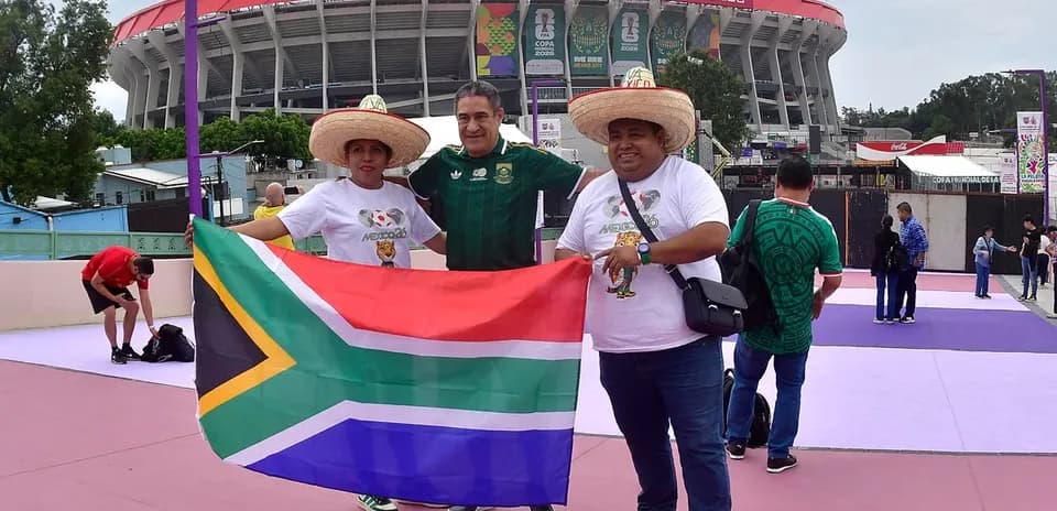Tres aficionados sosteniendo bandera de Sudáfrica frente al estadio
