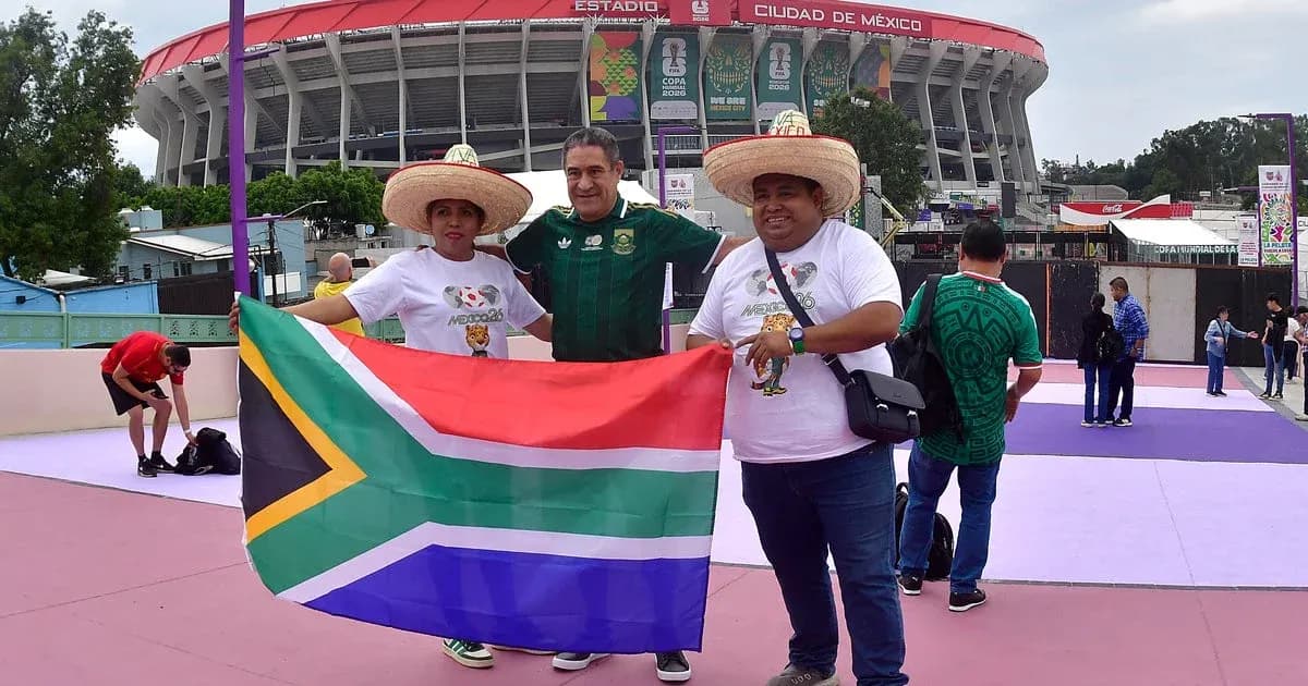Tres aficionados sosteniendo bandera de Sudáfrica frente al estadio